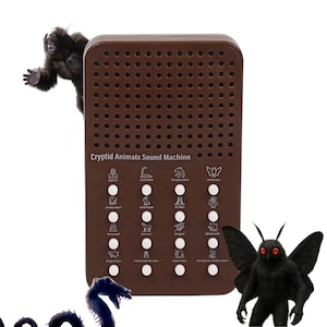 Op de afbeelding: Een bruine Cryptid Animals Sound Machine met een rooster van luidsprekergaten en knoppen. De machine bevat illustraties van cryptiden zoals Bigfoot, Mothman en de Jersey Devil. Een Bigfoot-figuur en een Mothman-figuur zijn bij de machine geplaatst.