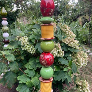 Könnte beinhalten: Eine vertikale Gartentotem-Skulptur bestehend aus gestapelten Keramikelementen, darunter eine rote Spitze, grüne Kugeln, gelbe gerippte Zylinder und flache runde Scheiben. Die Skulptur ist in einem wiederkehrenden Muster aus Farben und Formen angeordnet.