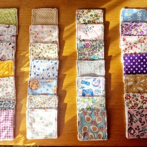 Puede incluir: Una colección de discos faciales reutilizables de tela cuadrada en varios patrones y colores. Los diseños incluyen flores, lunares, aguacates y mandos de videojuegos. Los discos están dispuestos en columnas ordenadas sobre una superficie de madera.