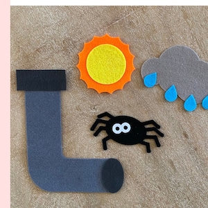 May include: Felt craft pieces for a children's learning game. The pieces include a sun, a cloud with raindrops, a spider, and a grey pipe.
