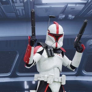 Könnte beinhalten: Eine weiße und rote Star Wars Klonkrieger-Actionfigur mit einem schwarzen Blaster in jeder Hand. Die Figur trägt einen weißen Helm mit einem roten Streifen und einem schwarzen Visier. Die Figur steht auf einem grau-schwarzen Hintergrund.