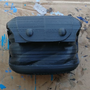 K&ouml;nnte beinhalten: Eine schwarze, 3D-gedruckte, erweiterbare, Rei&szlig;verschlusstasche mit einer Klappe und zwei Metalldruckkn&ouml;pfen.