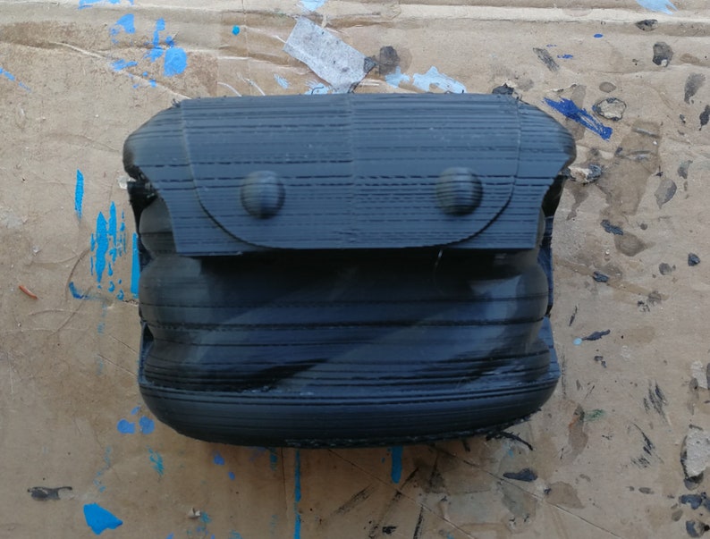 K&ouml;nnte beinhalten: Eine schwarze, 3D-gedruckte, erweiterbare, Rei&szlig;verschlusstasche mit einer Klappe und zwei Metalldruckkn&ouml;pfen.