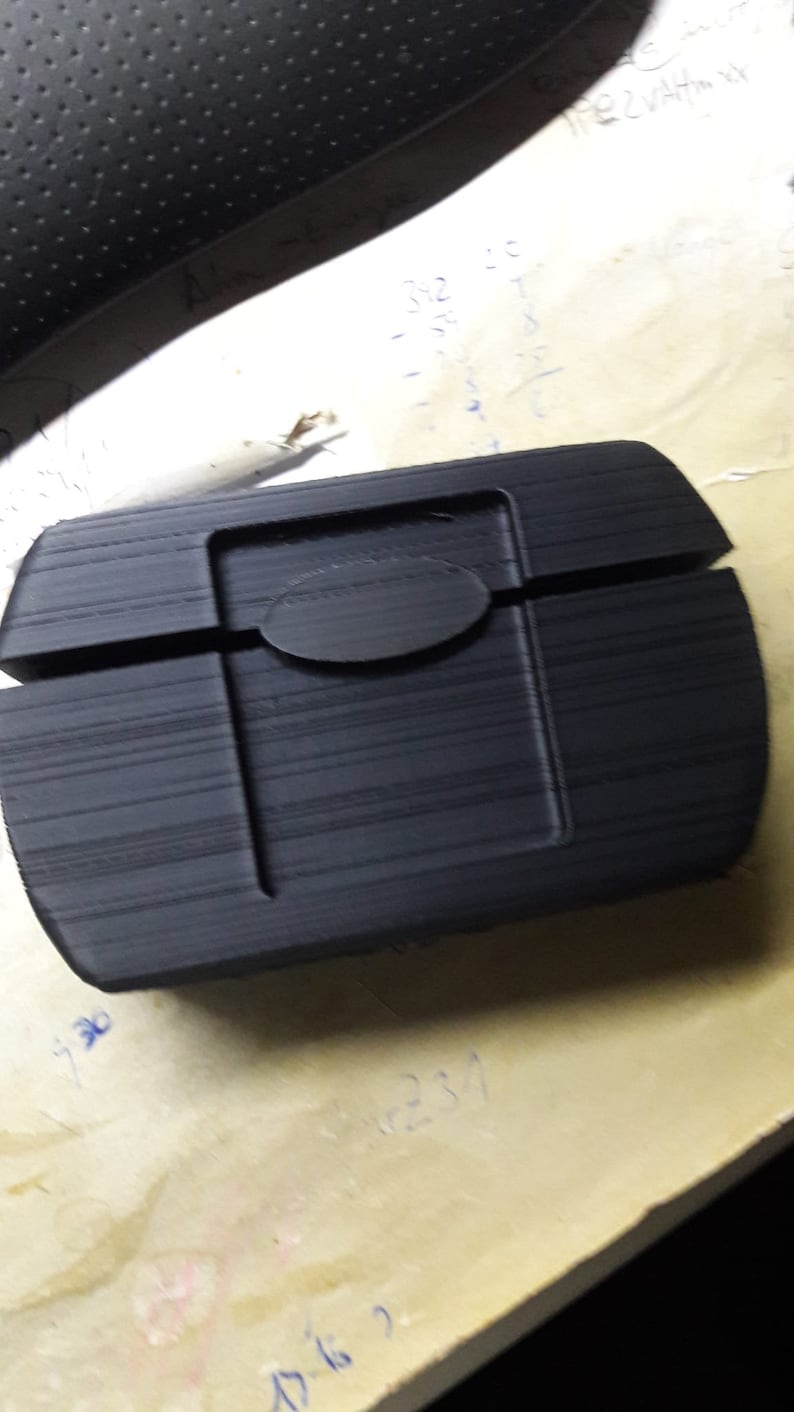 K&ouml;nnte beinhalten: Ein schwarzes, rechteckiges Plastiketui mit abgerundeter Kante und einem runden Ausschnitt oben. Das Etui hat eine Holzmaserung.