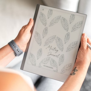 May include: A white e-reader with a gray border, displaying a page with a gray leaf pattern and the text "Meeting Journal" in black. The text "©/™ Remarkable" is at the bottom of the page.