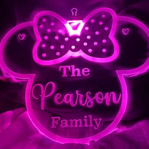 Op de afbeelding: Een roze verlicht Mickey Mouse-silhouet met een strik en stippen. De tekst "The Pearson Family" is onder de strik gegraveerd.