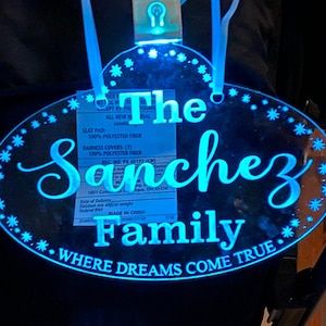 Op de afbeelding: Een blauw, ovaalvormig, verlicht bord met de tekst "The Sanchez Family" en de zin "Where Dreams Come True" in wit. Het bord is versierd met kleine witte sneeuwvlokken rond de rand.