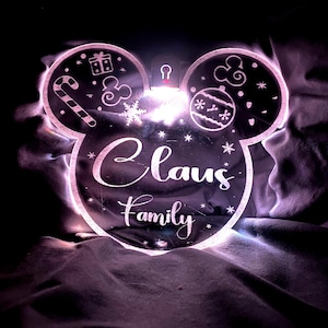 Op de afbeelding: Een wit Mickey Mouse-vormig verlicht bord met de woorden "Claus Family" en kerstthema-ontwerpen.