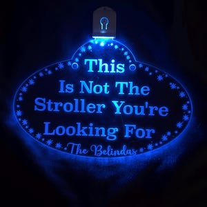 Op de afbeelding: Een blauw LED-bord met de tekst "This Is Not The Stroller You're Looking For" en "The Belindas" in een decoratieve ovale vorm. Het bord heeft een ophangbevestiging aan de bovenkant en is omgeven door stervormige lichten.