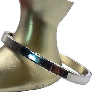May include: A silver-colored bangle bracelet with a square profile. The bracelet is displayed on a gold mannequin. The metal reflects light, creating highlights and shadows. The overall design is simple and modern.
