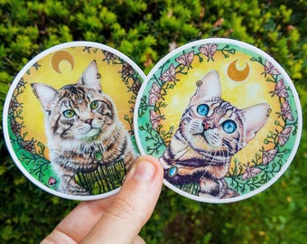 May include: Two round stickers featuring watercolor illustrations of cats with green eyes and a crescent moon. The cats are surrounded by floral designs and butterflies.