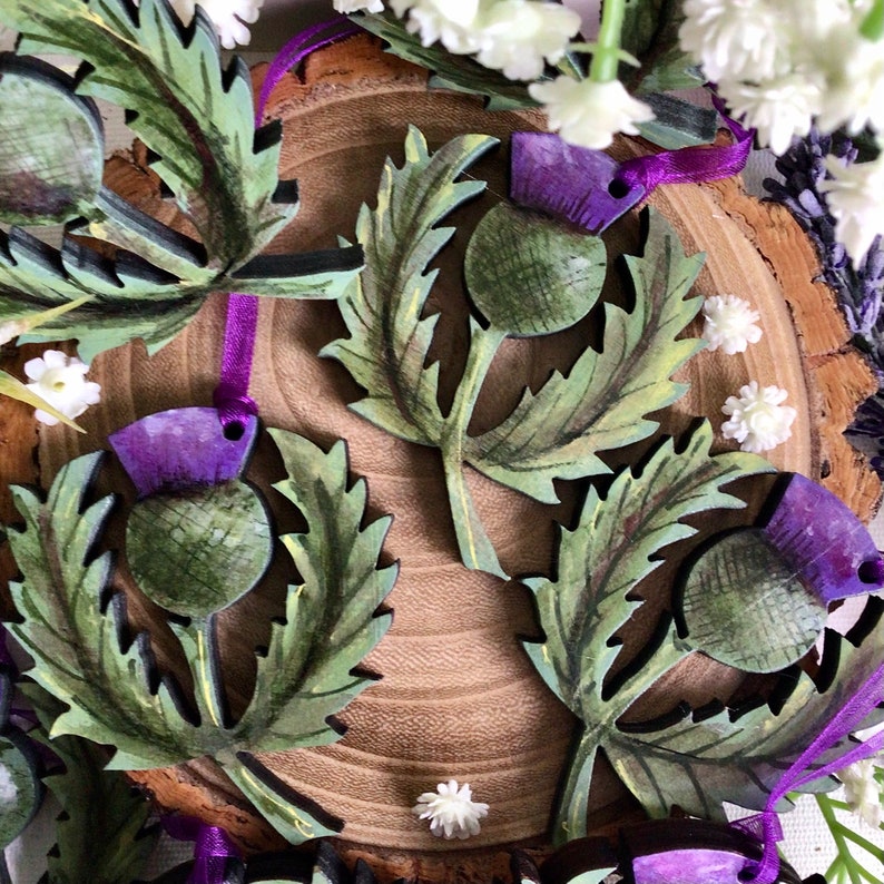 May include: A set of four wooden ornaments featuring hand-painted Scottish thistle designs. The thistles are green and purple with detailed leaves and stems. The ornaments are perfect for adding a touch of Scottish charm to your home decor.