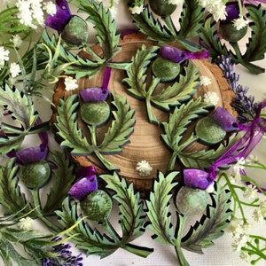 May include: A collection of wooden ornaments shaped like thistles with green leaves and purple flowers. The ornaments are painted in a realistic style and have a rustic charm.