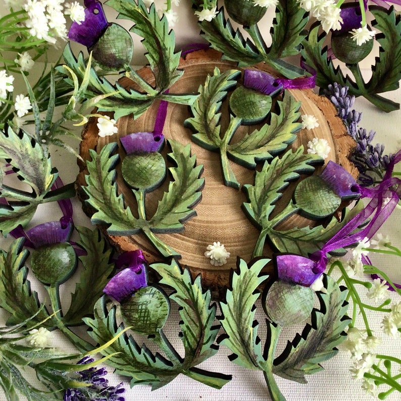 May include: A collection of wooden ornaments shaped like thistles with green leaves and purple flowers. The ornaments are painted in a realistic style and have a rustic charm.