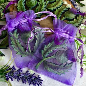 May include: Two purple organza bags with a green and purple painted thistle design. The bags are tied with purple ribbon and are sitting on a white surface with lavender flowers.
