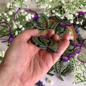 May include: A close-up of a hand holding a green and purple wooden thistle ornament. The ornament is surrounded by other thistle ornaments and white flowers.