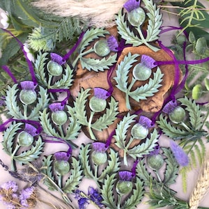 May include: A collection of wooden ornaments featuring a stylised Scottish thistle design. The ornaments are painted in shades of green and purple, with a realistic depiction of the thistle flower and leaves. Each ornament has a purple ribbon attached for hanging.