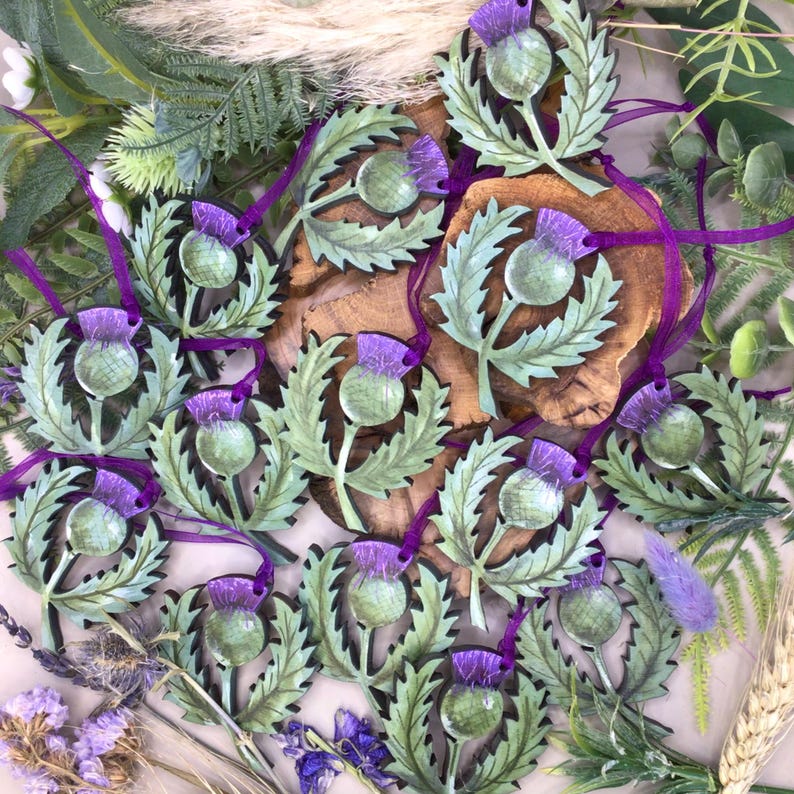 May include: A collection of wooden ornaments featuring a stylised Scottish thistle design. The ornaments are painted in shades of green and purple, with a realistic depiction of the thistle flower and leaves. Each ornament has a purple ribbon attached for hanging.