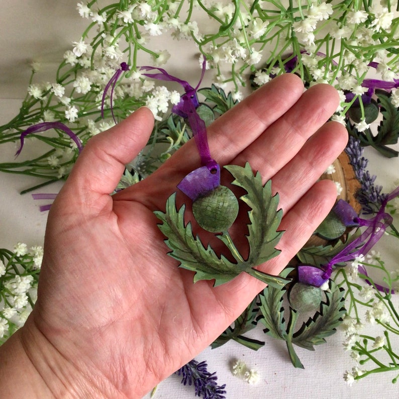 May include: A hand holding a wooden ornament shaped like a thistle, with green leaves and a purple flower. The ornament is painted with realistic details and has a purple ribbon attached.