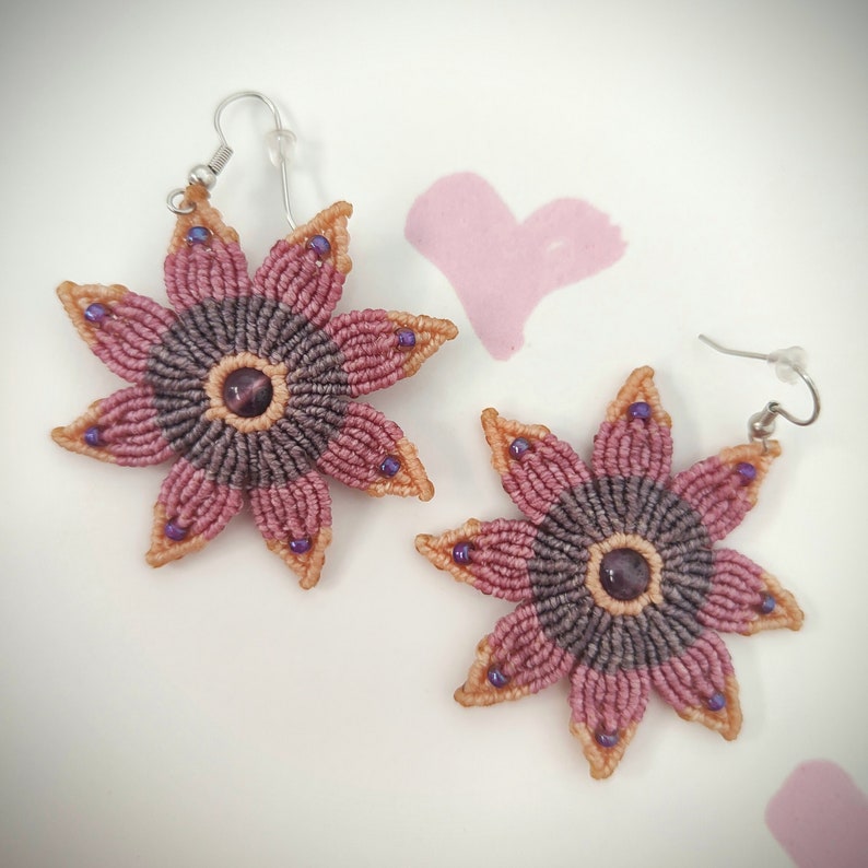 Puede incluir: Un par de pendientes hechos a mano con un dise&ntilde;o de flor con un centro morado y marr&oacute;n y un borde exterior de color melocot&oacute;n. Los pendientes tienen un gancho plateado.