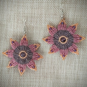 Puede incluir: Un par de pendientes con forma de flor con un efecto degradado rosa y morado. Los pendientes est&aacute;n hechos con una t&eacute;cnica de macram&eacute; y tienen una peque&ntilde;a piedra preciosa en el centro.