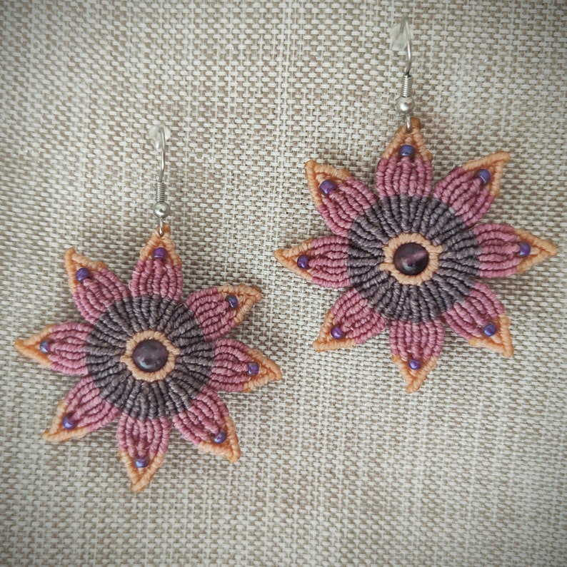 Puede incluir: Un par de pendientes con forma de flor con un efecto degradado rosa y morado. Los pendientes est&aacute;n hechos con una t&eacute;cnica de macram&eacute; y tienen una peque&ntilde;a piedra preciosa en el centro.