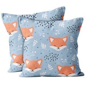 May include: Two square cushions with a light blue background featuring a repeating pattern of orange fox faces, white leaves, black speckles, and blue dots. The cushions are designed for home decor.