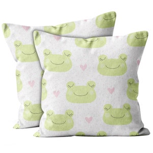 May include: Two white pillowcases with a repeating pattern of green frogs with closed eyes and pink hearts. The pillowcases are made of a soft, textured fabric.