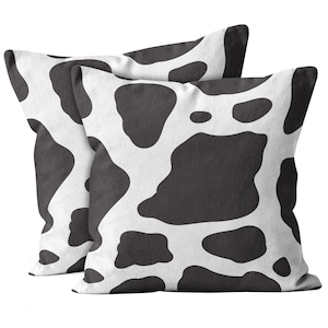 May include: Two black and white cow print throw pillows. The pillows are made of a soft, textured fabric.