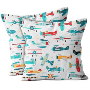 May include: Two square white cushions featuring a repeating pattern of colourful aeroplanes. The planes are in various colours, including red, blue, yellow, and green, with propeller details and star accents. The cushions appear to be made of a soft fabric.