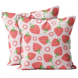 May include: Two square cushions featuring a pattern of red strawberries with white seeds and green stalks, and pink flowers on a pale pink backdrop. The cushions are made of a soft material.