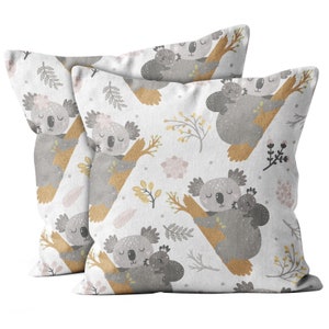 May include: Two square throw pillows with a repeating pattern of gray koalas, each holding a baby koala, on a white background. The design includes tan branches, pink flowers, and gray and yellow leaves.