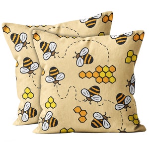 May include: Two square throw pillows with a light tan background. The pillows feature a repeating pattern of cartoon bees, honeycombs, and dotted flight paths in black, yellow, and orange. The pillows are likely made of a soft fabric.