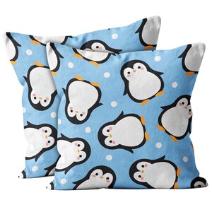 May include: Two square throw pillows with a light blue background covered in cartoon penguins and white polka dots. The penguins have black bodies, white bellies, orange feet, and pink cheeks. The pillows are a decorative home accessory.