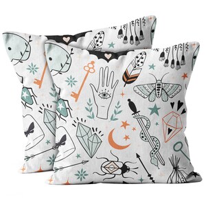 May include: Two white throw pillows with a repeating pattern of mystical symbols. The design includes keys, hands with an eye, feathers, moths, crystals, arrows, and crescent moons in orange, teal, and black.