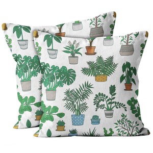 Pode incluir: Duas almofadas quadradas com fundo branco e um padrão repetido de plantas em vasos de várias formas e tamanhos. As plantas são em tons de verde, com vasos cinza, marrom e azul. As almofadas têm cantos de madeira.
