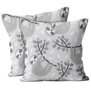 May include: Two square throw pillows with a repeating pattern of gray sloths hanging from tree branches with leaves on a light gray background. The sloths have smiling faces and are in various poses.