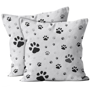 May include: Two white throw pillows featuring a black paw print design. The pillows are made from a soft, comfortable fabric and are ideal for adding a touch of whimsy to any room.