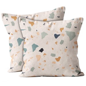 May include: Two square throw pillows with a terrazzo pattern. The pillows have a cream-colored background with scattered shapes in shades of blue, tan, and black. The pillows are made of a soft fabric and are likely used for home decor.