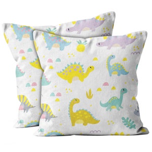 May include: Two square cushions featuring a white background adorned with a repeating pattern of colourful cartoon dinosaurs. The dinosaurs are yellow, blue, and purple, with pink, yellow, and green accents. The cushions are likely made of soft fabric.