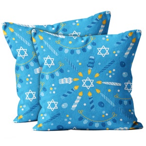 May include: Two square throw pillows with a blue background and a repeating pattern of white Stars of David, candles, and dreidels. The candles have yellow flames, and the dreidels are blue and white.