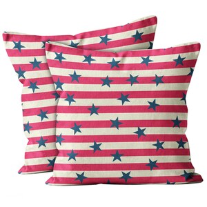May include: Two square cushions with a patriotic design. The cushions feature alternating red and off-white stripes, with navy blue stars scattered across the surface. The cushions are made of a soft fabric and are likely used for home decor.
