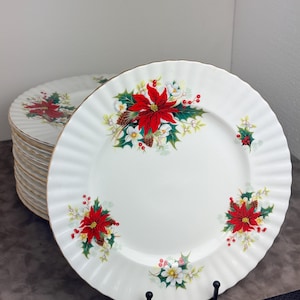 May include: A stack of white ceramic dinner plates with a scalloped edge and a gold-tone rim. Each plate features a repeating holiday pattern of red poinsettia flowers, green holly leaves, small white flowers, and red berries around the border.
