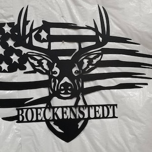 May include: Black metal silhouette of a deer head with antlers, set against a distressed American flag design. The name "BOECKENStedt" is displayed below the deer head.