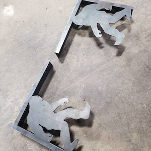 May include: Two metal shelf brackets shaped like a silhouette of a Bigfoot. The brackets are in a corner shape and are made of metal.