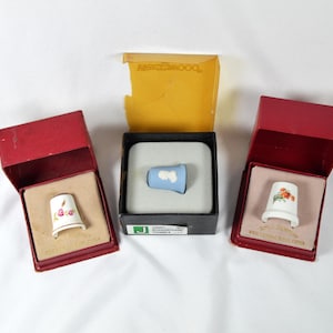 May include: Three decorative thimbles in their original boxes. One thimble is light blue with a white cloud design, and the other two are white with floral patterns. The thimbles are displayed in red and black boxes.