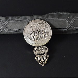 May include: A silver-toned metal brooch with a detailed relief scene of figures gathered outdoors. The brooch has an ornate, scroll-like design below the circular scene. The background is black, with a gray fabric element.