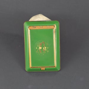 May include: A rectangular, green metal compact with gold trim. The compact has a hinged lid and a gold emblem on the front. The background is a dark gray surface.