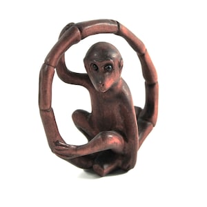 May include: A brown monkey figurine seated within a segmented ring. The monkey has a detailed face and is posed with its hands resting on its legs. The ring appears to be made of the same material as the monkey, and the background is white.