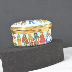 May include: An oval-shaped light blue enamel trinket box with a gold-tone metal rim. The exterior features a colorful illustration of various figures in traditional clothing holding hands, with the large gold-colored text 2000 printed on the front.